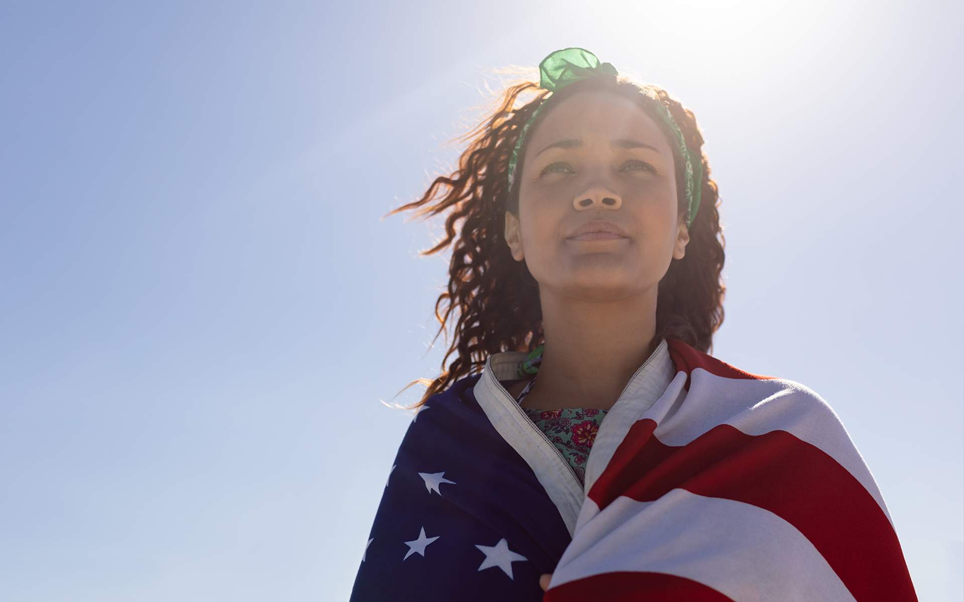 Photo of a young Black woman with an American flag wrapped around her shoulders. She is standing in the sun with her eyes closed, looking calm and determined.