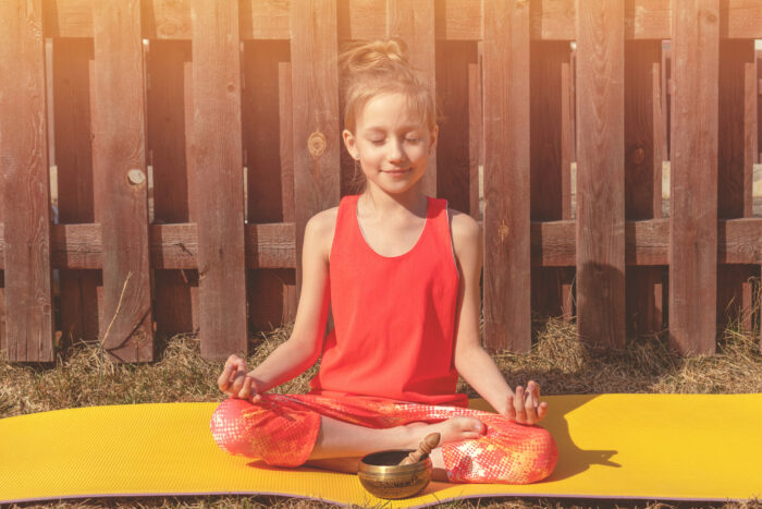 A teenager girl in an orange tracksuit is meditating in the courtyard of her house. The sun is shining. Singing bowl for meditation