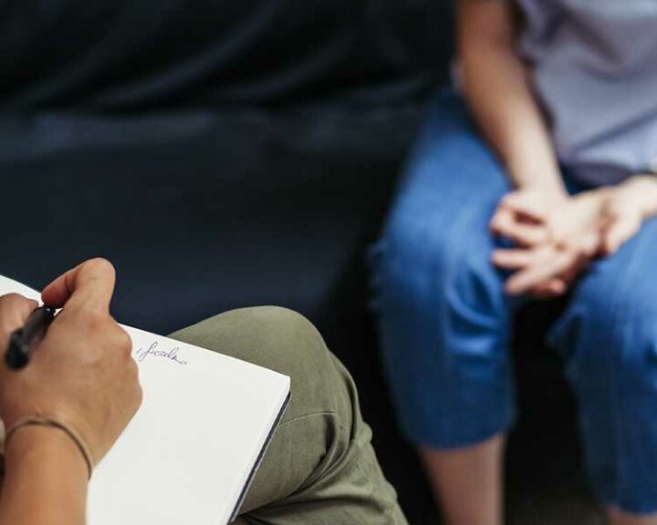 A therapist takes notes while client faces them on a couch wringing their hands