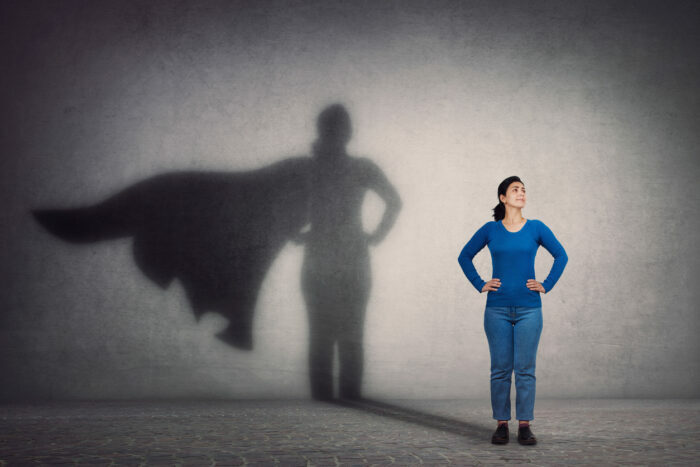 Brave woman keeps arms on hips, smiling confident, casting a superhero with cape shadow on the wall