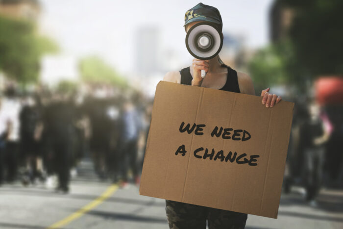 public protest and riots in the city streets. woman with megaphoen and cardboard poster in hands says "we need a change" and protesters crowd in the background