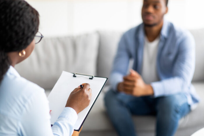 Young guy sitting on a couch having a session with a psychotherapist in an office, the psychotherapist has a clip board in hand and is writing something on it