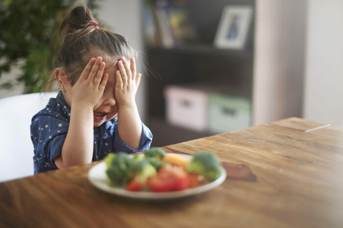 child covering their eyes with their hands while sitting in front of a plate of vegetables