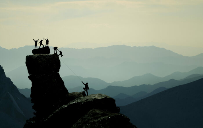 group of people standing on a mountain high fiving and helping someone up while overlooking other mountains