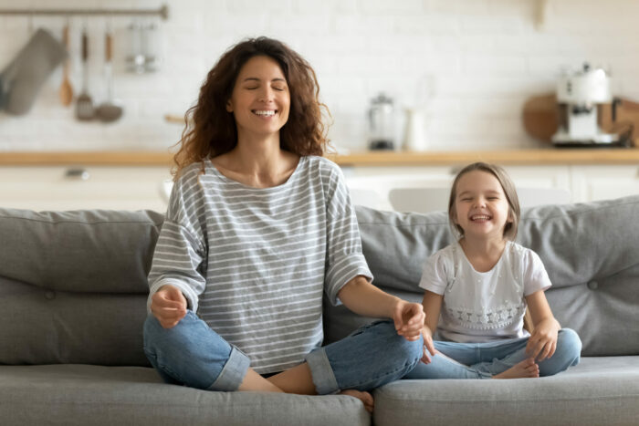 Happy mom teaching small daughter yoga breathing exercise on couch.